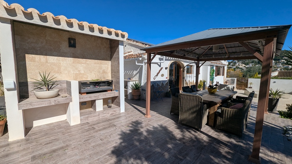 Zona de comedor exterior con barbacoa de obra revestida en piedra y una amplia pérgola que cubre una mesa con sillas de mimbre sobre suelo cerámico.