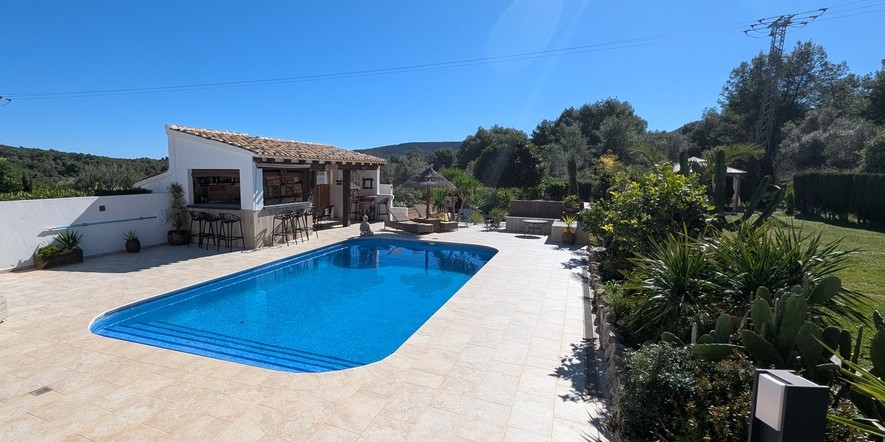Zona de piscina con terraza de piedra, cocina de verano equipada con barra y taburetes, rodeada de jardines mediterráneos y vistas despejadas al campo.