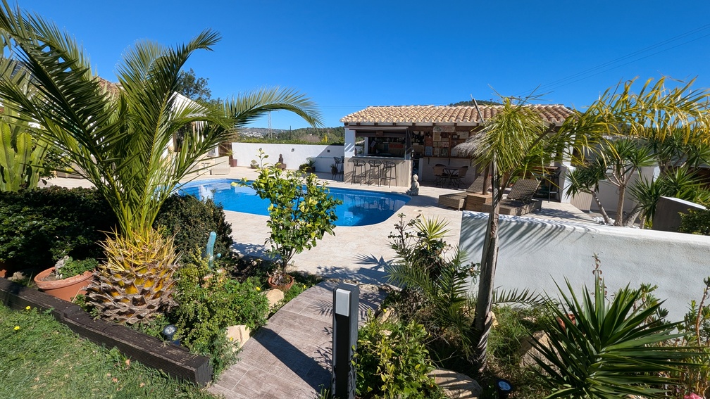 Zona de piscina con terraza de piedra, jardín mediterráneo con palmeras y una cocina de verano techada con barra y taburetes exteriores.