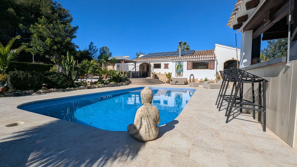 Zona de piscina privada con pavimentación de piedra, estatua de Buda, barra exterior con taburetes y vivienda con paneles solares fotovoltaicos.