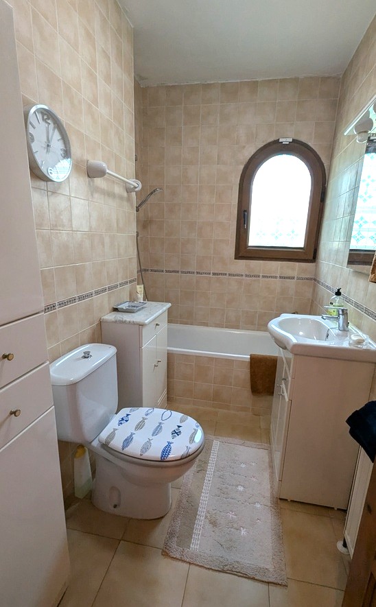 Baño de estilo tradicional con azulejos en tonos beige, bañera integrada y ventana de arco. Incluye mueble de lavabo blanco y armarios auxiliares para almacenamiento.