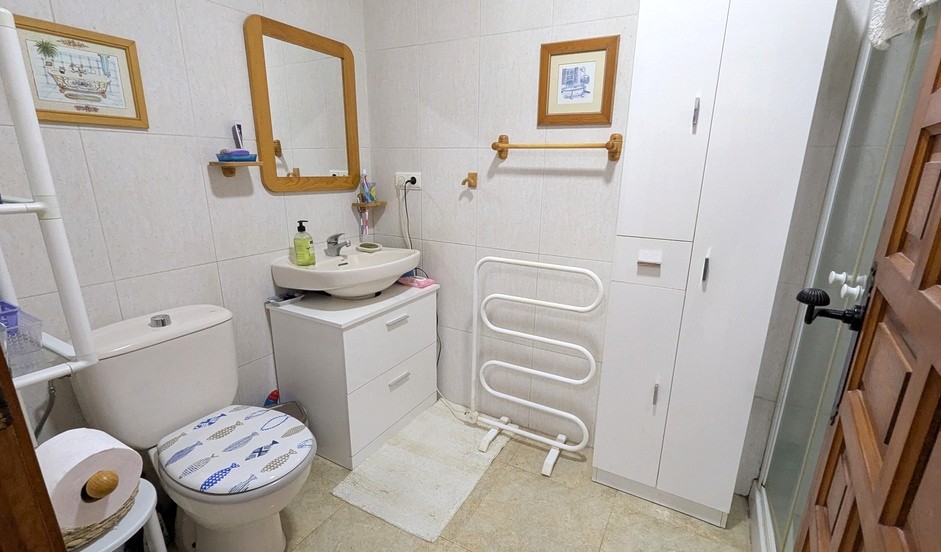 Cuarto de baño funcional con mueble de lavabo blanco, espejo con marco de madera y armarios de almacenaje. Incluye radiador toallero y paredes alicatadas.