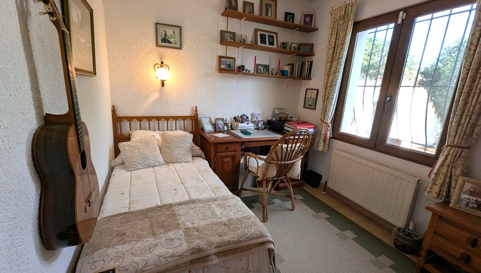 Dormitorio con cama individual de madera, escritorio clásico, silla de mimbre y estanterías. Cuenta con calefacción por radiador y ventana protegida con rejas.