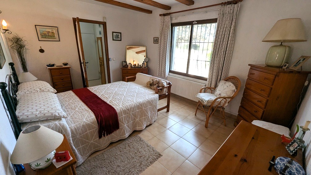 Dormitorio de estilo tradicional con vigas de madera vista, suelos de baldosa y calefacción por radiador bajo la ventana principal con luz natural.