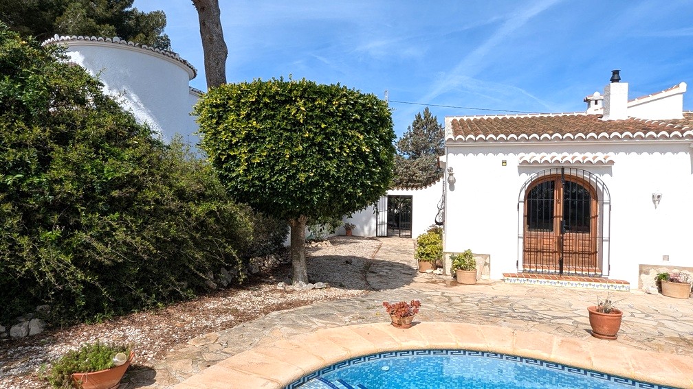 Exterior de la villa con piscina de mosaico, terraza de piedra natural y fachada blanca tradicional con puertas de madera en arco y rejas de hierro forjado.