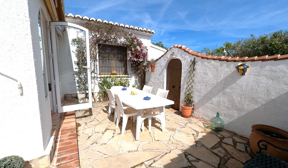 Patio exterior privado con suelo de piedra natural, conjunto de comedor blanco y arquitectura mediterránea tradicional con arcos y remates de teja cerámica.