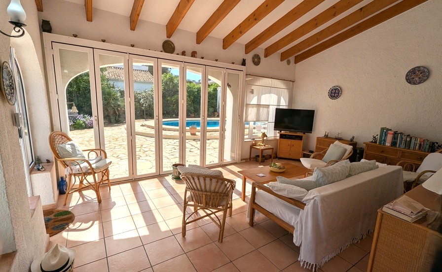 Salón luminoso con vigas de madera a la vista, suelo de terracota y amplias puertas plegables de cristal con acceso directo a la terraza y piscina.