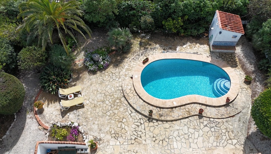 Vista elevada de la piscina de forma arriñonada con gresite azul, rodeada por una amplia terraza de piedra natural y un jardín mediterráneo con palmeras y vegetación madura.