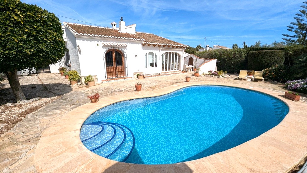 Vista exterior de la piscina de forma arriñonada con gresite azul, rodeada de una terraza de piedra y la fachada blanca de estilo tradicional mediterráneo.