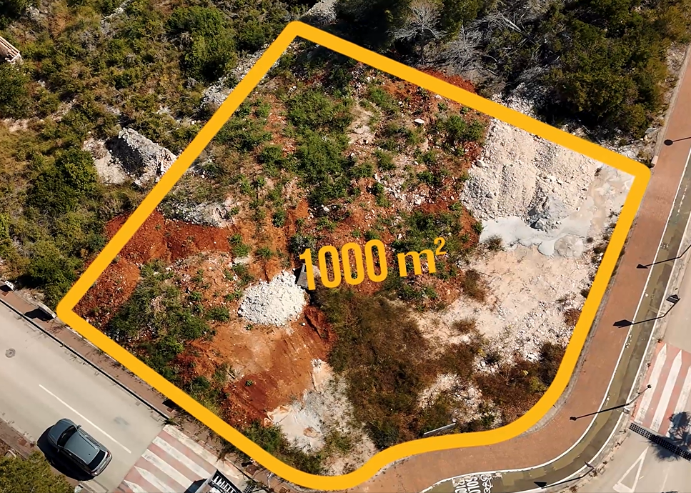 Vista aérea de la parcela de 1.000 m² en Jávea, destacando su ubicación en esquina, el terreno de tierra rojiza y la infraestructura urbana circundante con aceras.