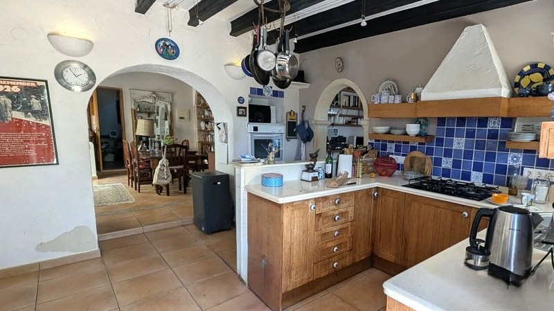 Cocina tradicional con armarios de madera, salpicadero de azulejos azules y vigas oscuras. Arcos de estilo Tosca conectan con el comedor y suelos de barro cocido.
