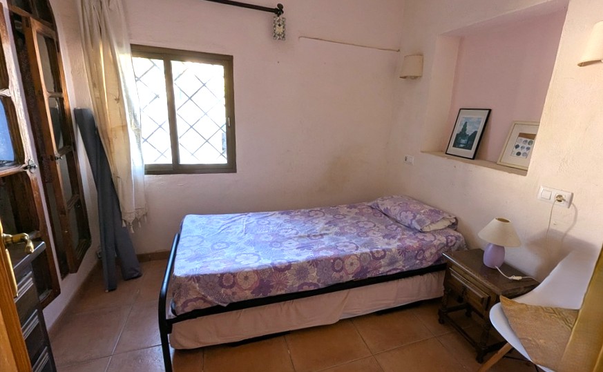 Dormitorio acogedor con cama individual, suelos de terracota y una hornacina decorativa en la pared. Incluye una ventana con rejas tradicionales y luz natural.