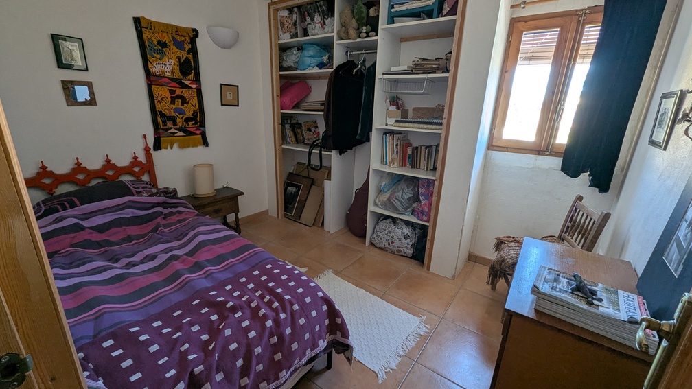 Dormitorio con suelos de baldosa de barro, estanterías abiertas integradas y ventana de madera. Incluye una cama individual y escritorio de madera bajo luz natural.