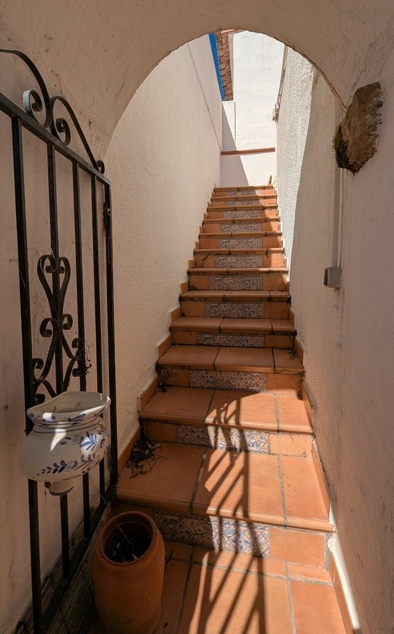 Escalera exterior con peldaños de terracota y contrahuellas de azulejo decorativo, situada tras un arco blanco con cancela de hierro forjado.