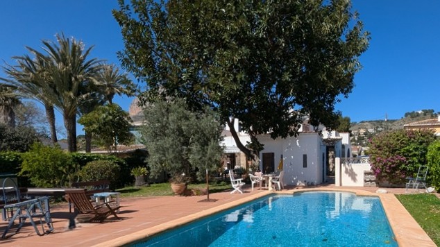 Piscina privada rodeada de una terraza de terracota y un jardín maduro con palmeras, ofreciendo vistas despejadas al Montgó y la fachada de la villa.