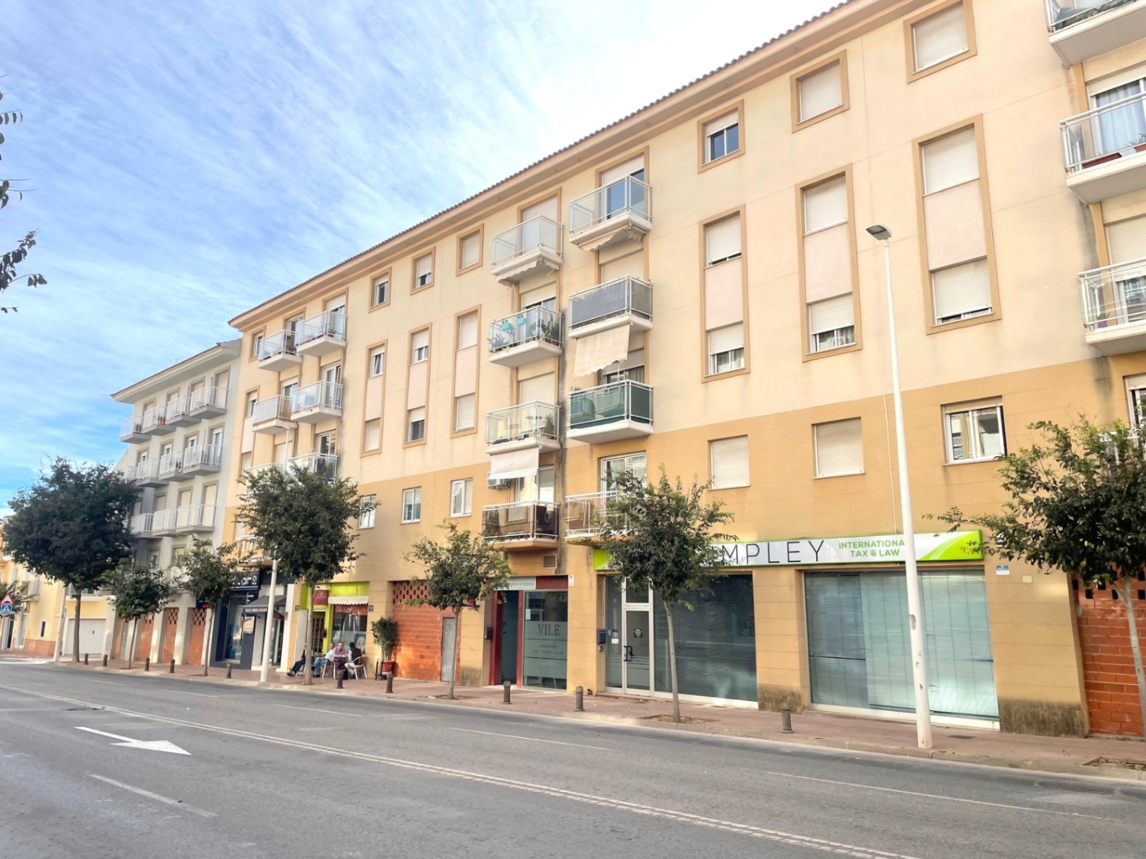 Fachada exterior de un edificio residencial con locales comerciales en planta baja, situado en una avenida principal con aceras arboladas y balcones acristalados.
