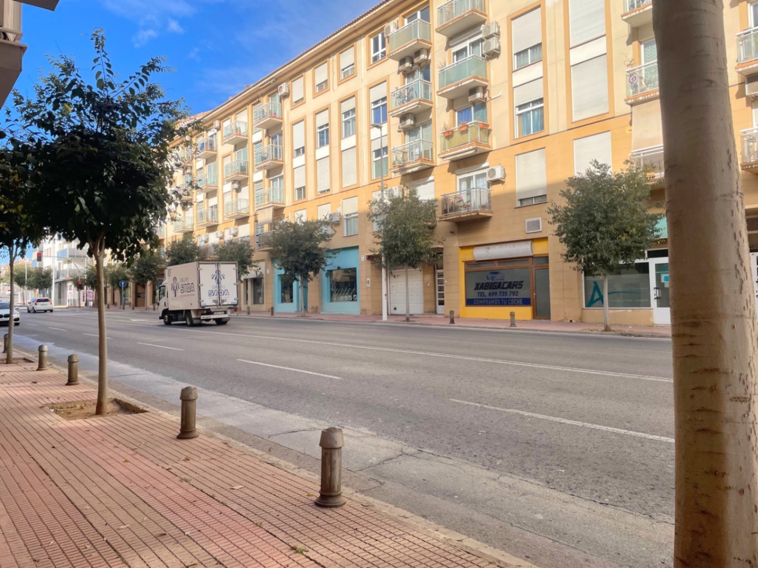 Vista exterior de la Avenida Palmela en Jávea, mostrando la fachada del edificio con locales comerciales en planta baja y amplias aceras arboladas.