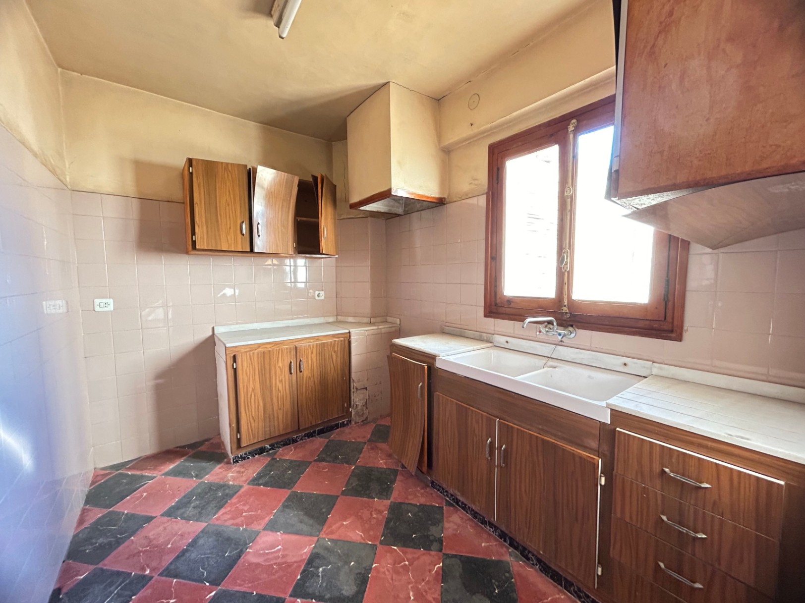 Cocina original con suelos de damero en tonos rojizos y negros, armarios de madera y encimera de mármol blanco. Dispone de fregadero de doble seno y ventana de madera.