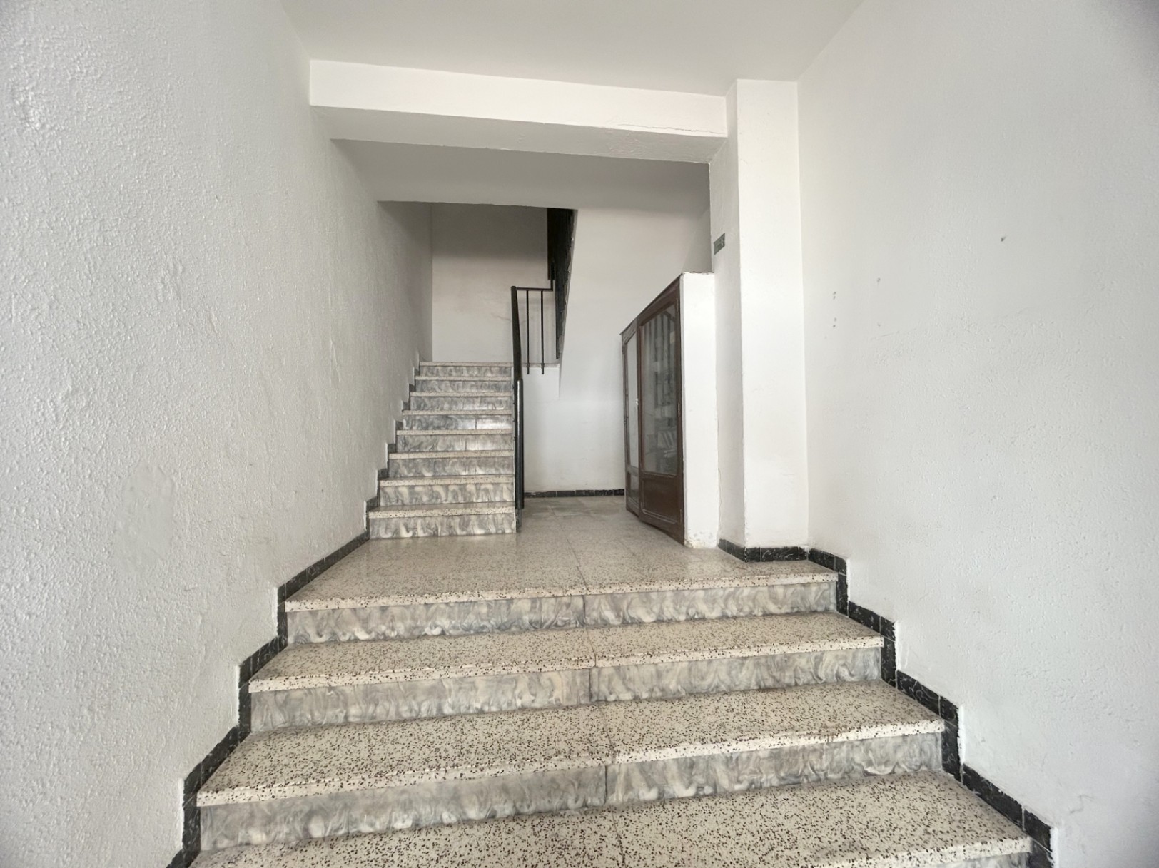 Escalera comunitaria con peldaños de terrazo y frontales jaspeados, paredes blancas y barandilla metálica negra. Incluye un armario de madera en el rellano intermedio.