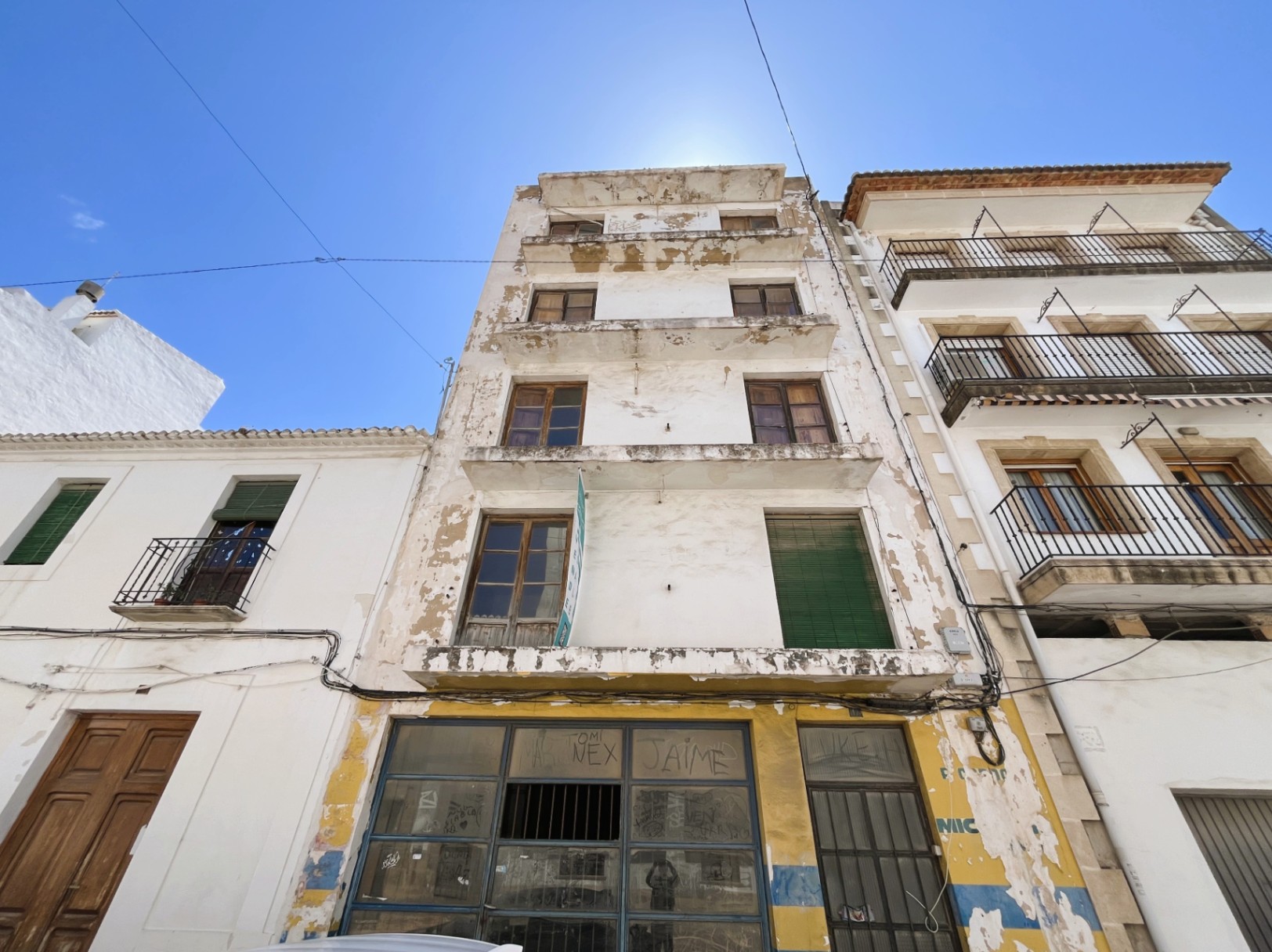 Fachada exterior de cuatro alturas con local comercial en planta baja. Presenta balcones corridos y carpintería de madera bajo un cielo despejado en el centro de Jávea.