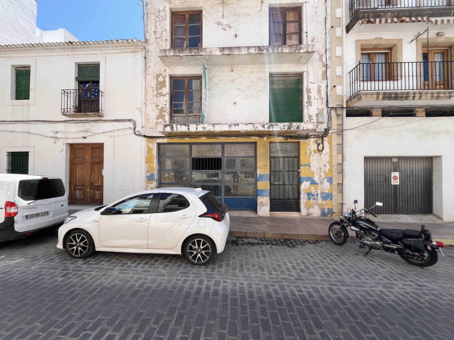 Fachada exterior del edificio en Jávea con local comercial en planta baja, balcones longitudinales y ventanas de madera sobre una calle de adoquines tradicional.