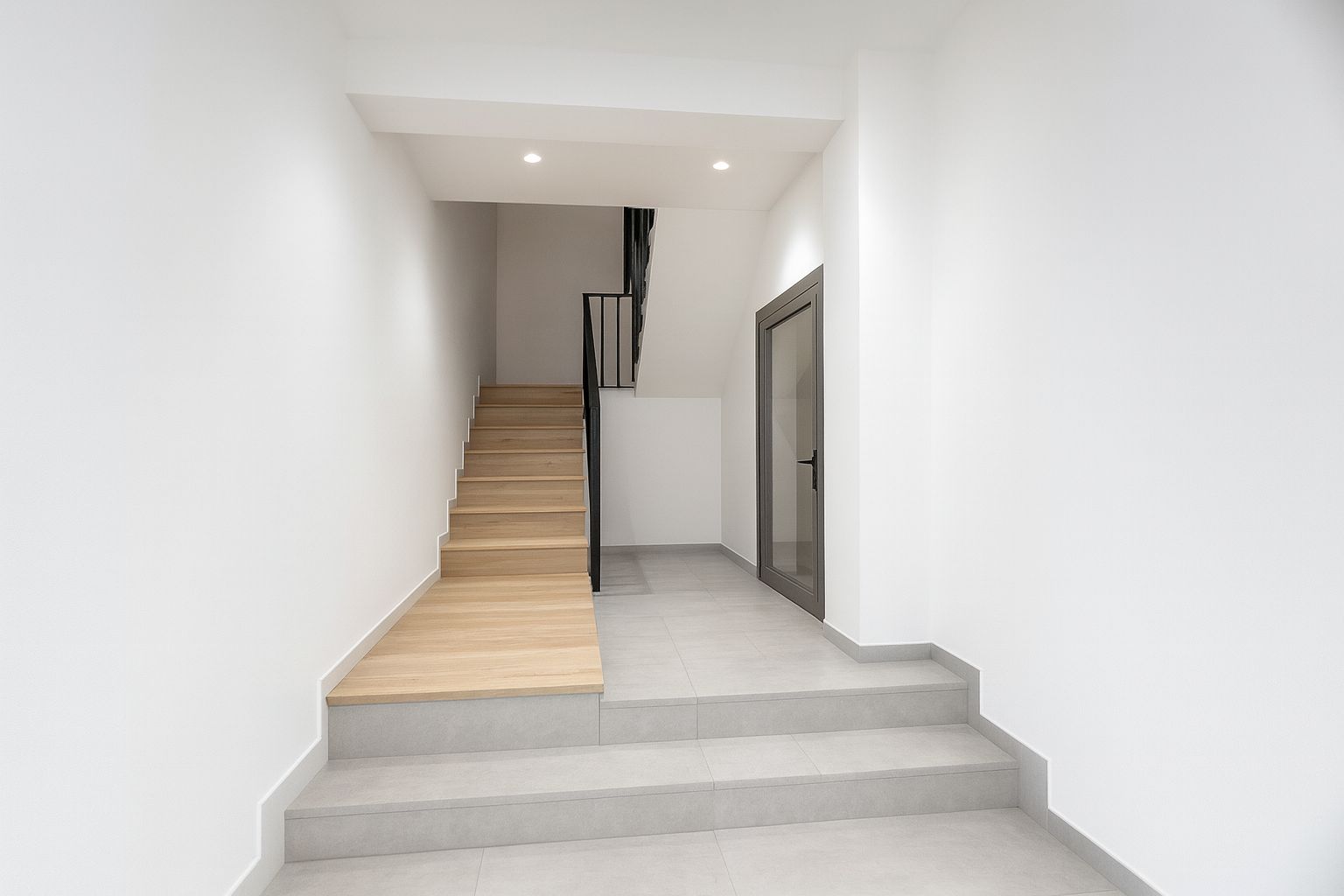 Vestíbulo de entrada con escalera de madera clara y barandilla metálica negra. Suelos de gres gris, paredes blancas y puerta acristalada con marco de aluminio.