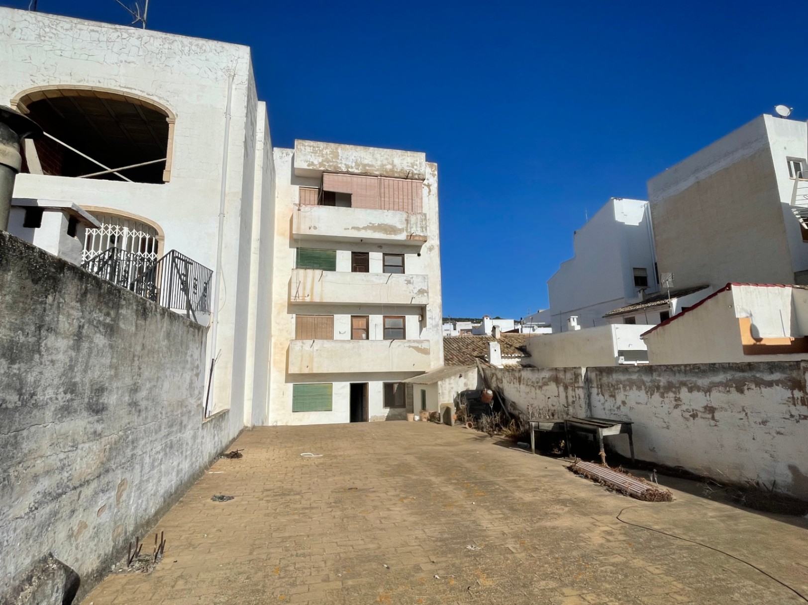 Vista posterior del edificio de cuatro plantas con un amplio patio pavimentado y muros perimetrales. La fachada muestra balcones y ventanales originales bajo un cielo despejado.