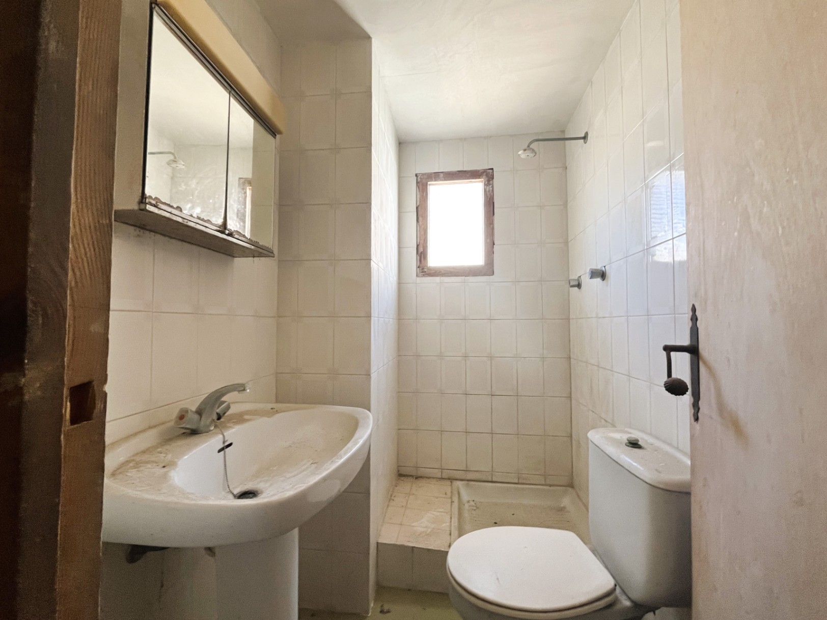 Baño funcional con azulejos blancos, lavabo de pedestal y zona de ducha con ventana pequeña. Un espacio con gran potencial de reforma integral.
