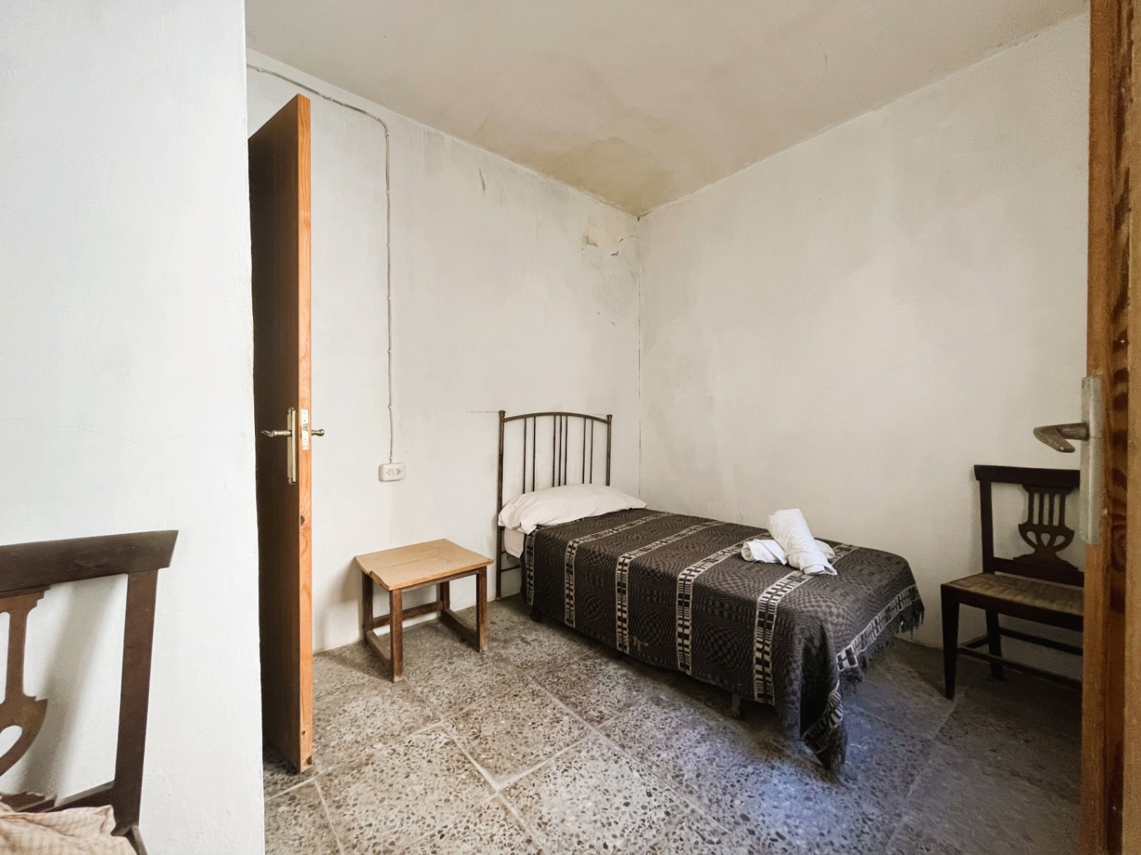 Dormitorio sencillo con suelos de terrazo original, cama individual con cabecero de forja y mobiliario de madera tradicional en una estancia de paredes blancas.