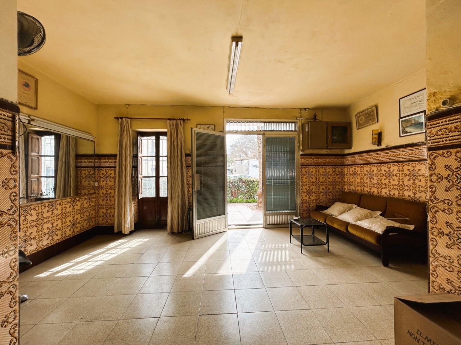 Amplia estancia de entrada con zócalos de azulejos tradicionales, suelos cerámicos y acceso directo al exterior a través de grandes ventanales que aportan luz natural.