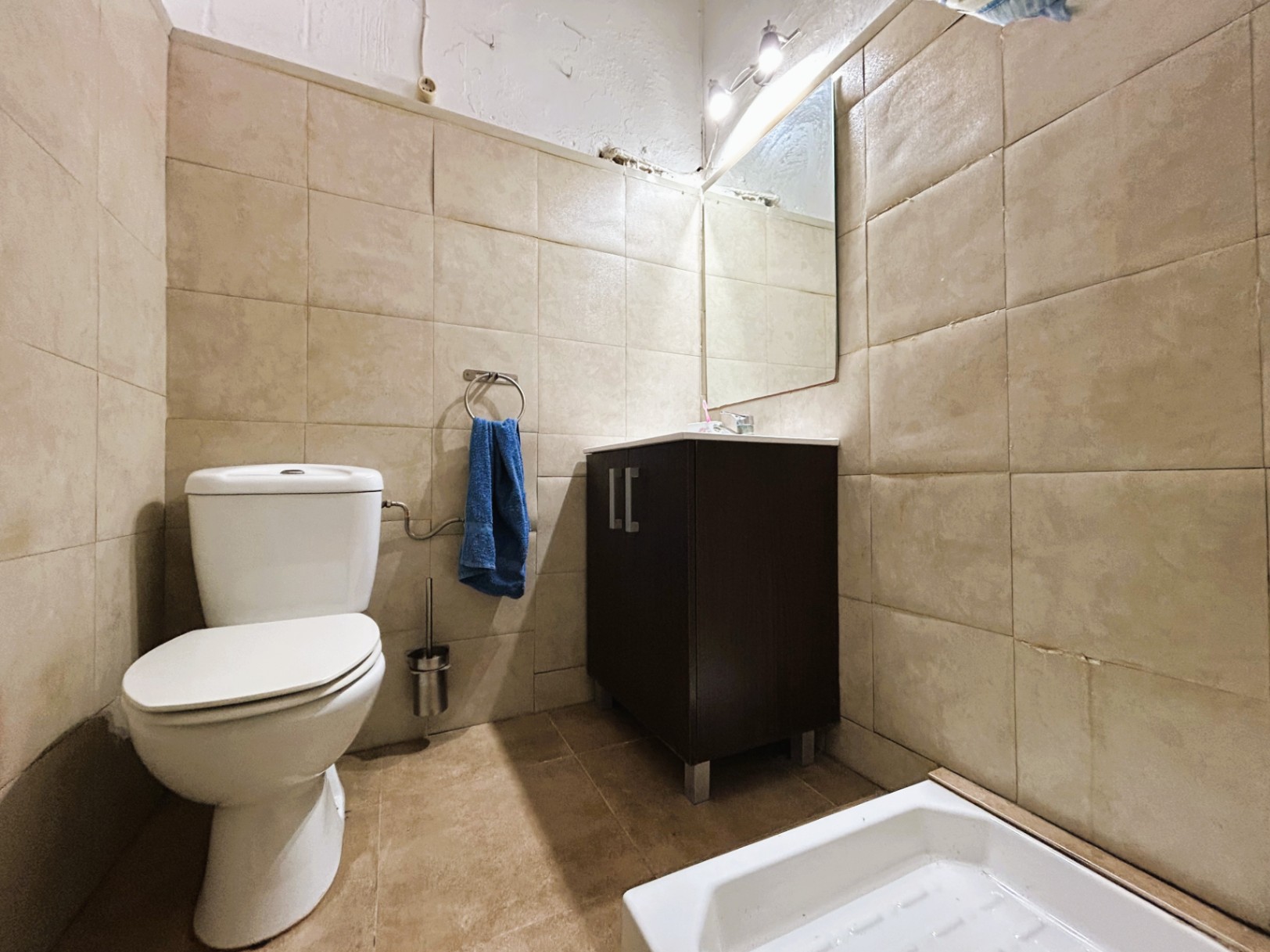 Cuarto de baño funcional con paredes alicatadas en tonos beige, inodoro cerámico blanco y mueble de lavabo en madera oscura con espejo iluminado.