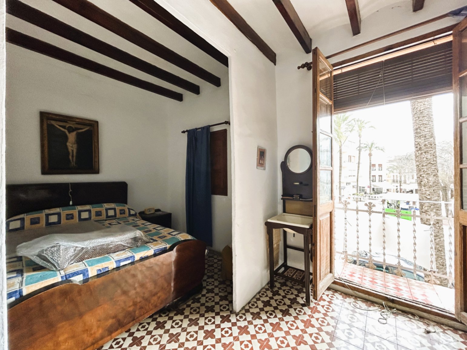 Dormitorio tradicional con vigas de madera vista, suelos de baldosa hidráulica y acceso directo a un balcón con vistas a la plaza y palmeras.