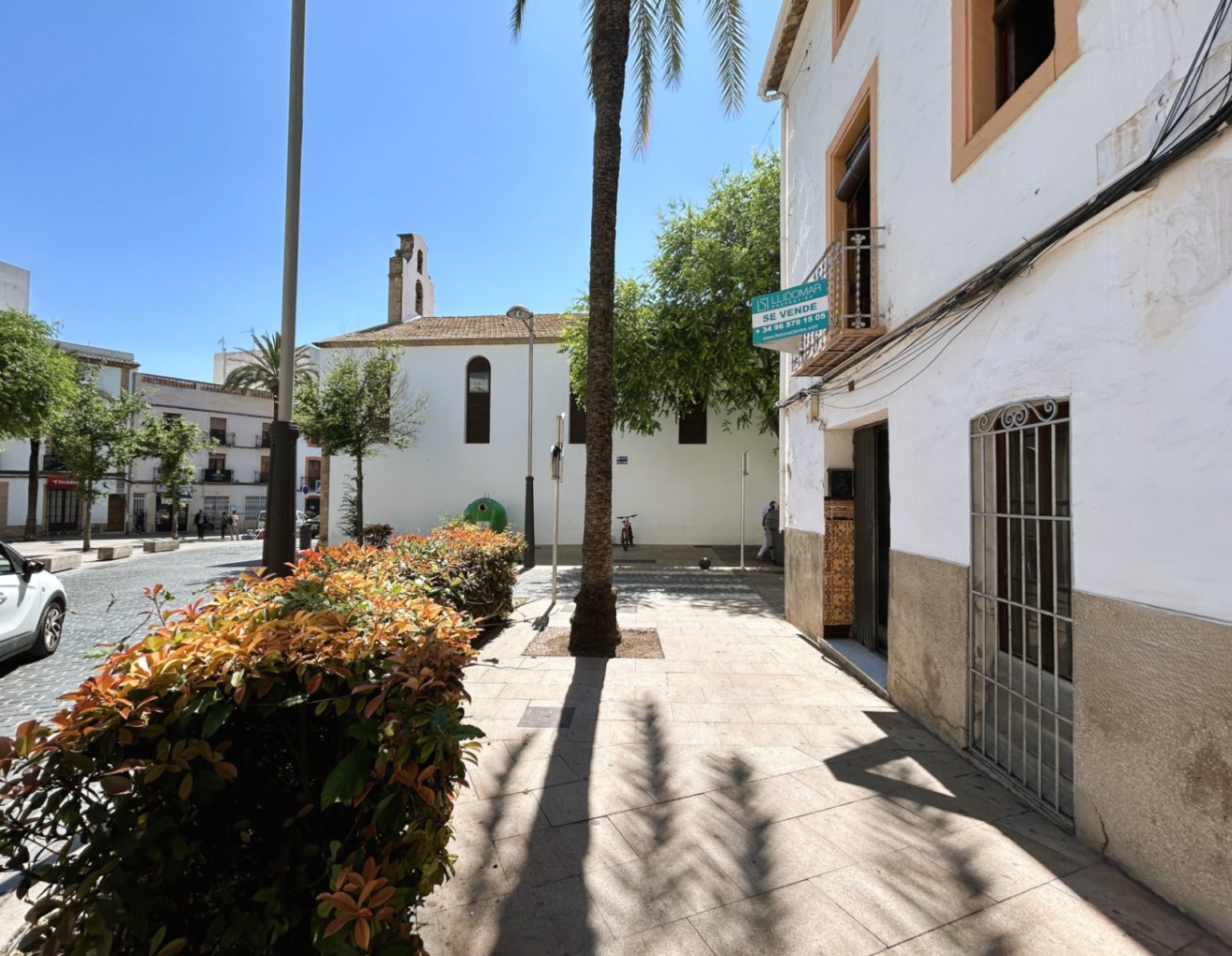 Fachada blanca con zócalo de piedra en el casco antiguo de Jávea, situada junto a una plaza peatonal con palmeras y vistas a un campanario histórico.