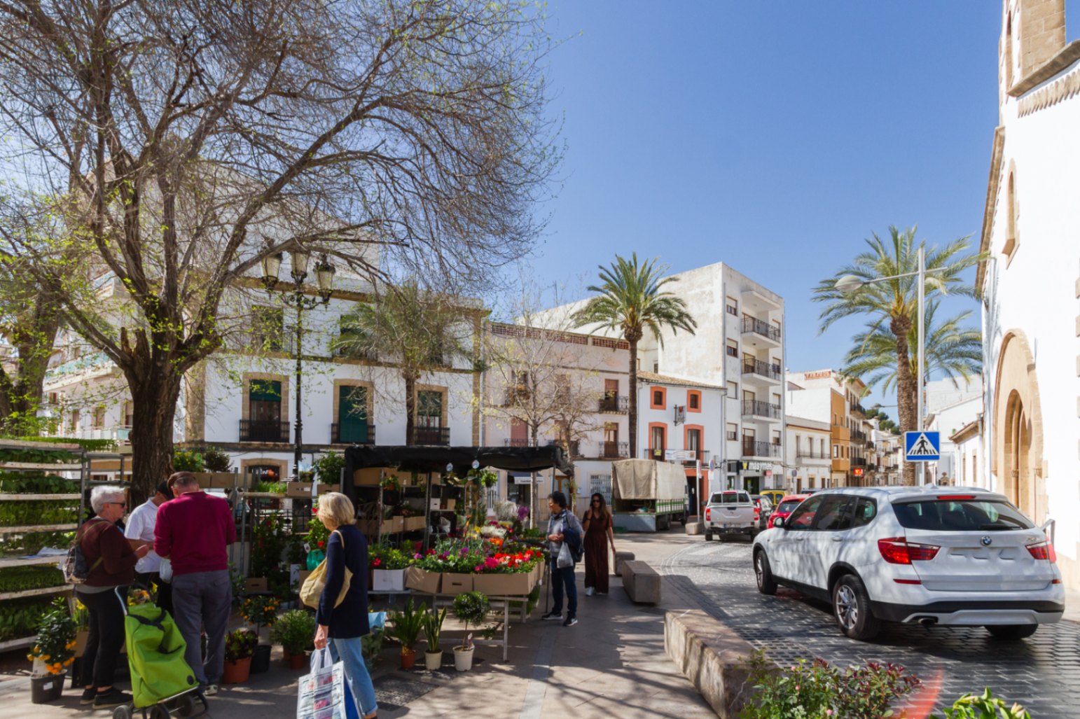 Vista de una plaza en el casco antiguo de Jávea con un mercado de flores, rodeada de arquitectura tradicional, calles empedradas y palmeras bajo luz natural intensa.