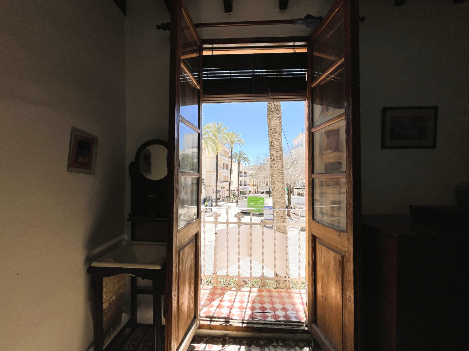 Vista desde el interior a través de puertas de madera hacia un balcón con suelo de damero, ofreciendo vistas despejadas a una plaza con palmeras en el casco antiguo.