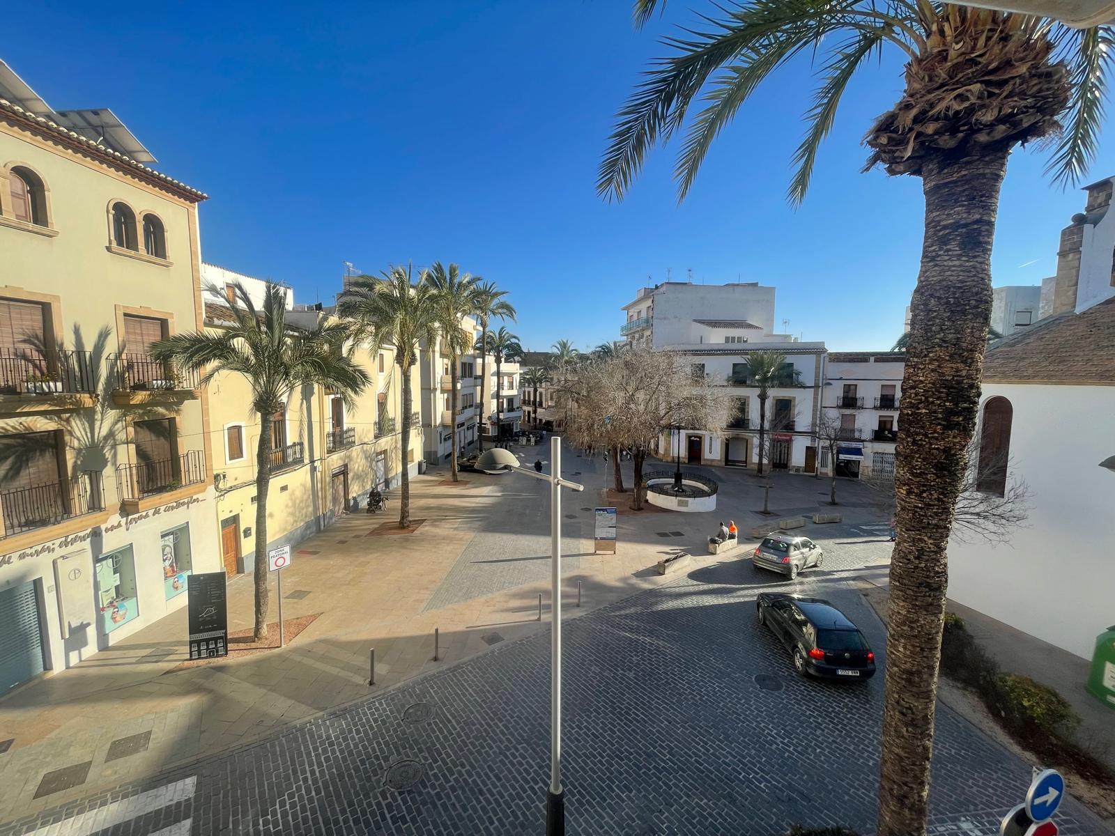 Vista elevada desde la propiedad hacia una plaza peatonal empedrada en el casco antiguo de Jávea, rodeada de arquitectura tradicional y palmeras.