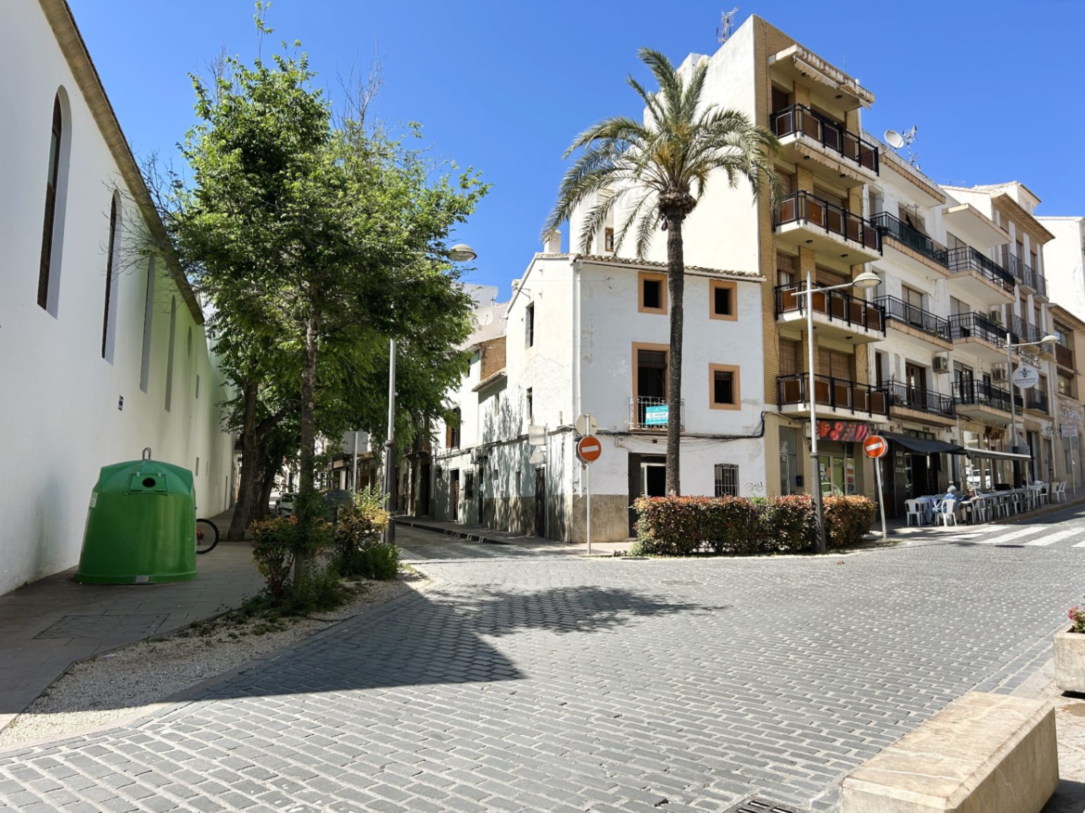 Vista exterior de la fachada blanca en esquina situada en una calle adoquinada del casco antiguo de Jávea, junto a una palmera y edificios tradicionales.