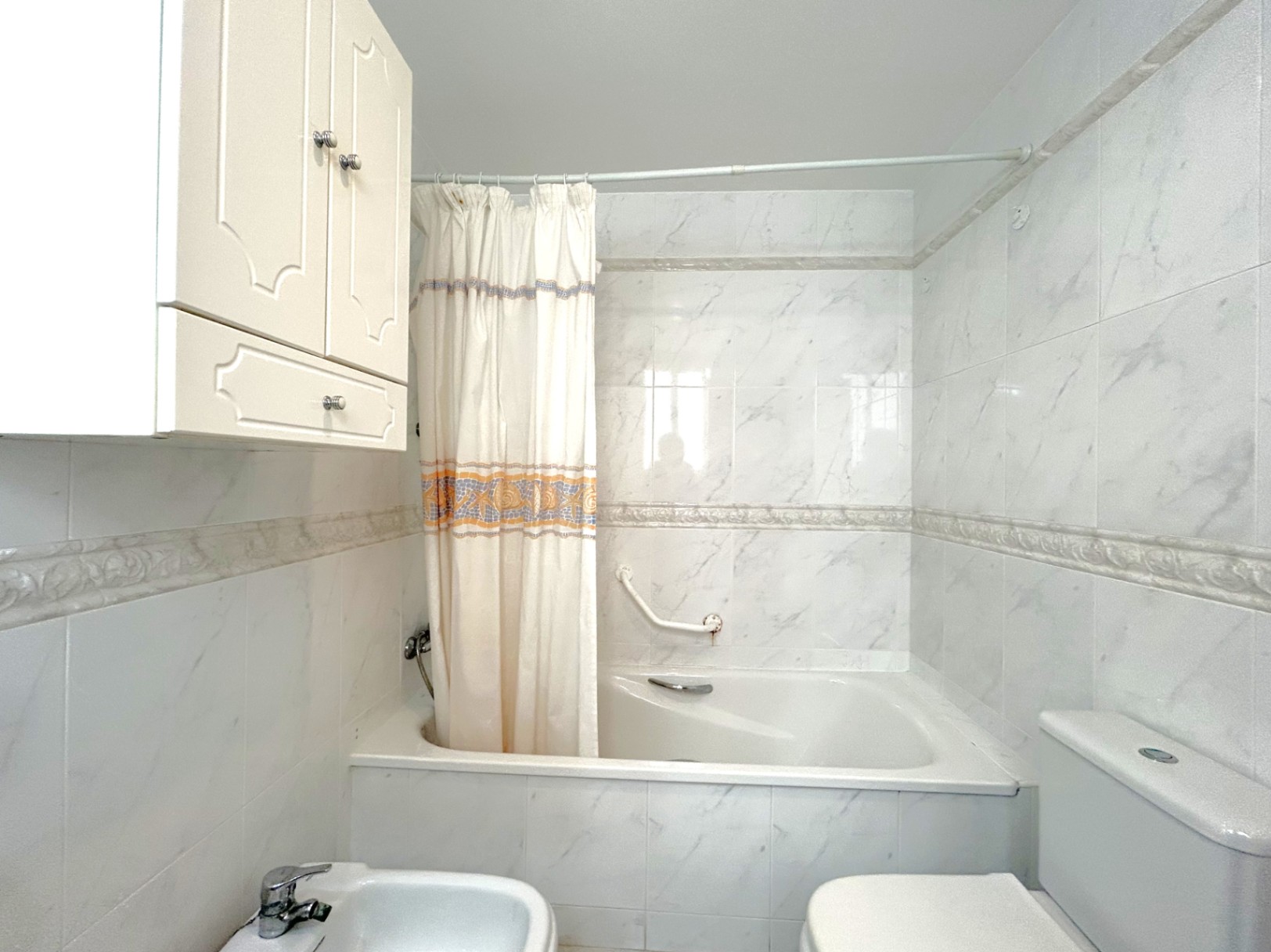 Baño con bañera integrada, alicatado de efecto mármol y cenefa decorativa. Incluye bidé, inodoro y un armario auxiliar de pared en color blanco.