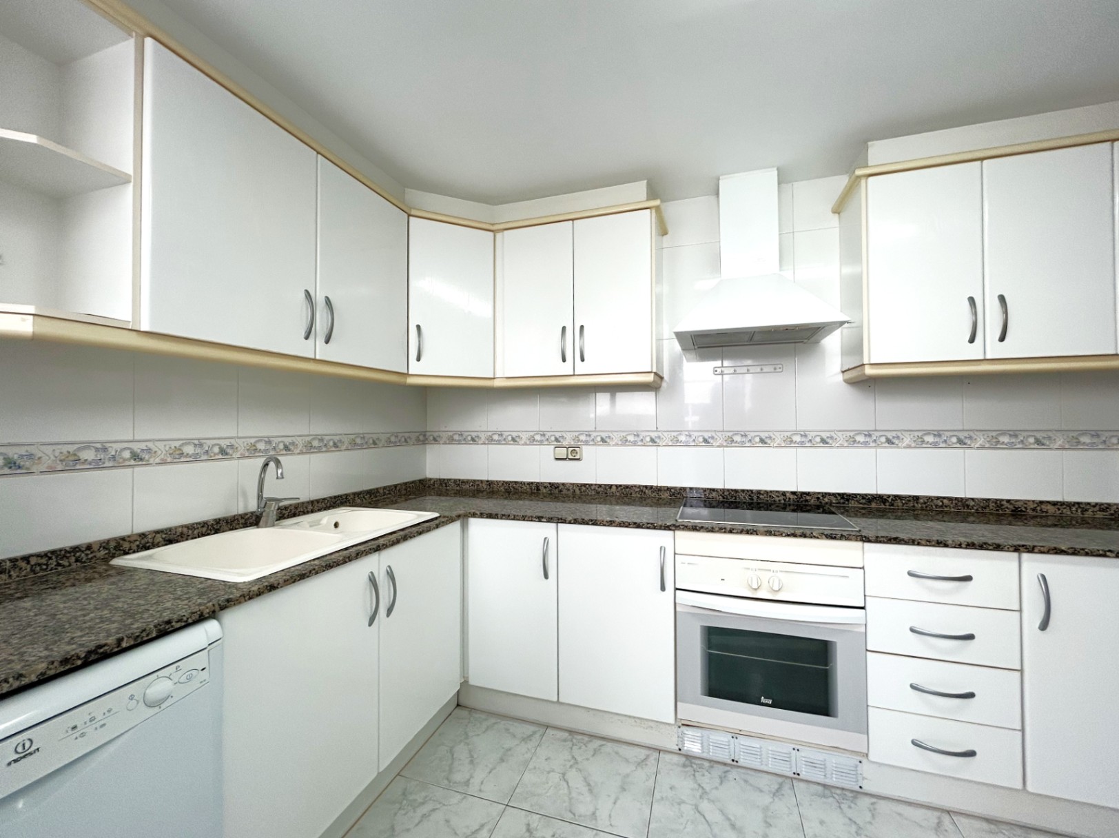 Cocina independiente equipada con armarios blancos, encimera de granito, placa vitrocerámica y horno. Incluye azulejos con cenefa decorativa y suelos de gres.