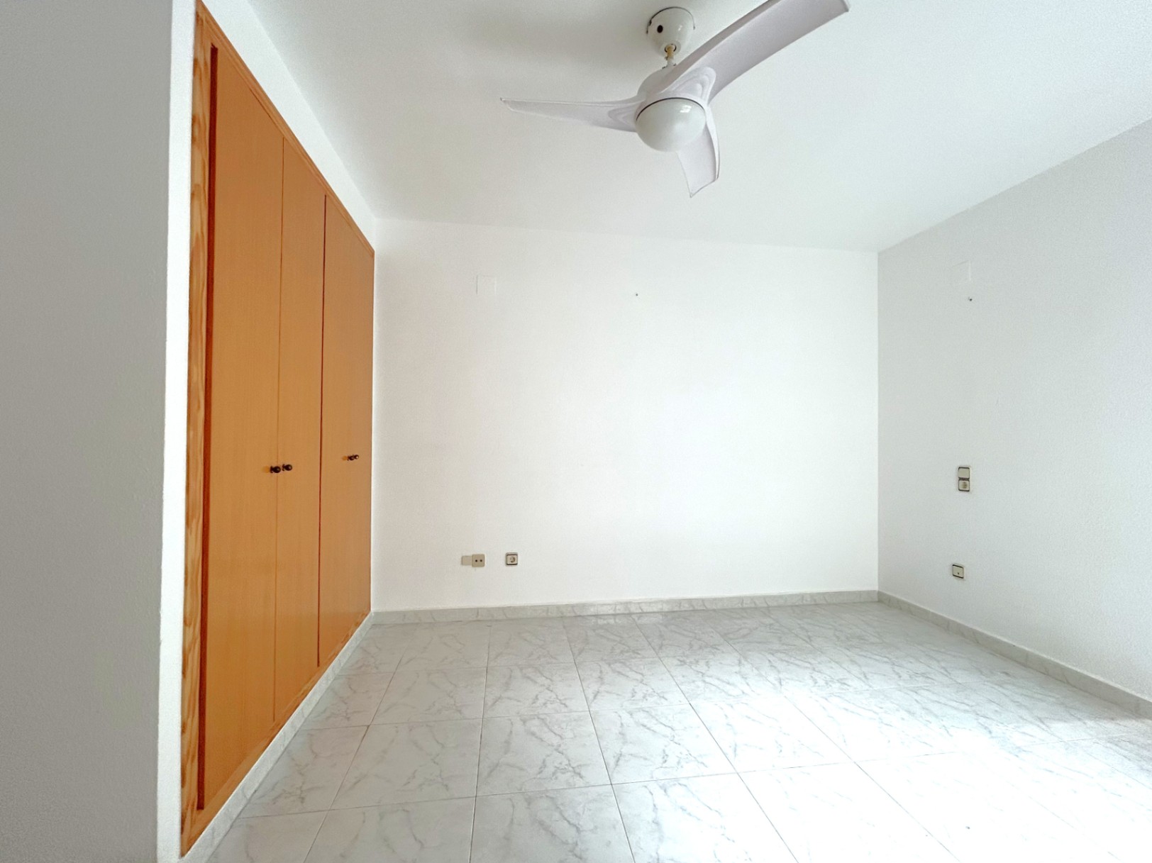 Dormitorio luminoso con armario empotrado de madera, suelos de gres con efecto mármol y ventilador de techo de tres aspas en blanco.
