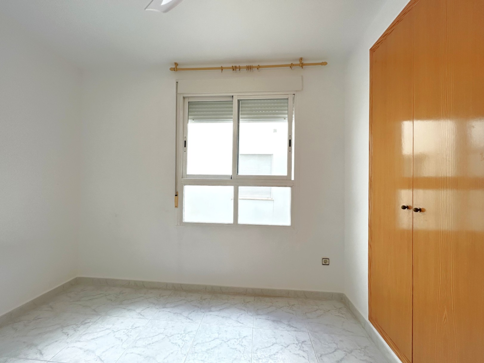 Dormitorio luminoso con suelos de gres efecto mármol, armario empotrado de madera y ventana de aluminio blanco con persiana integrada.