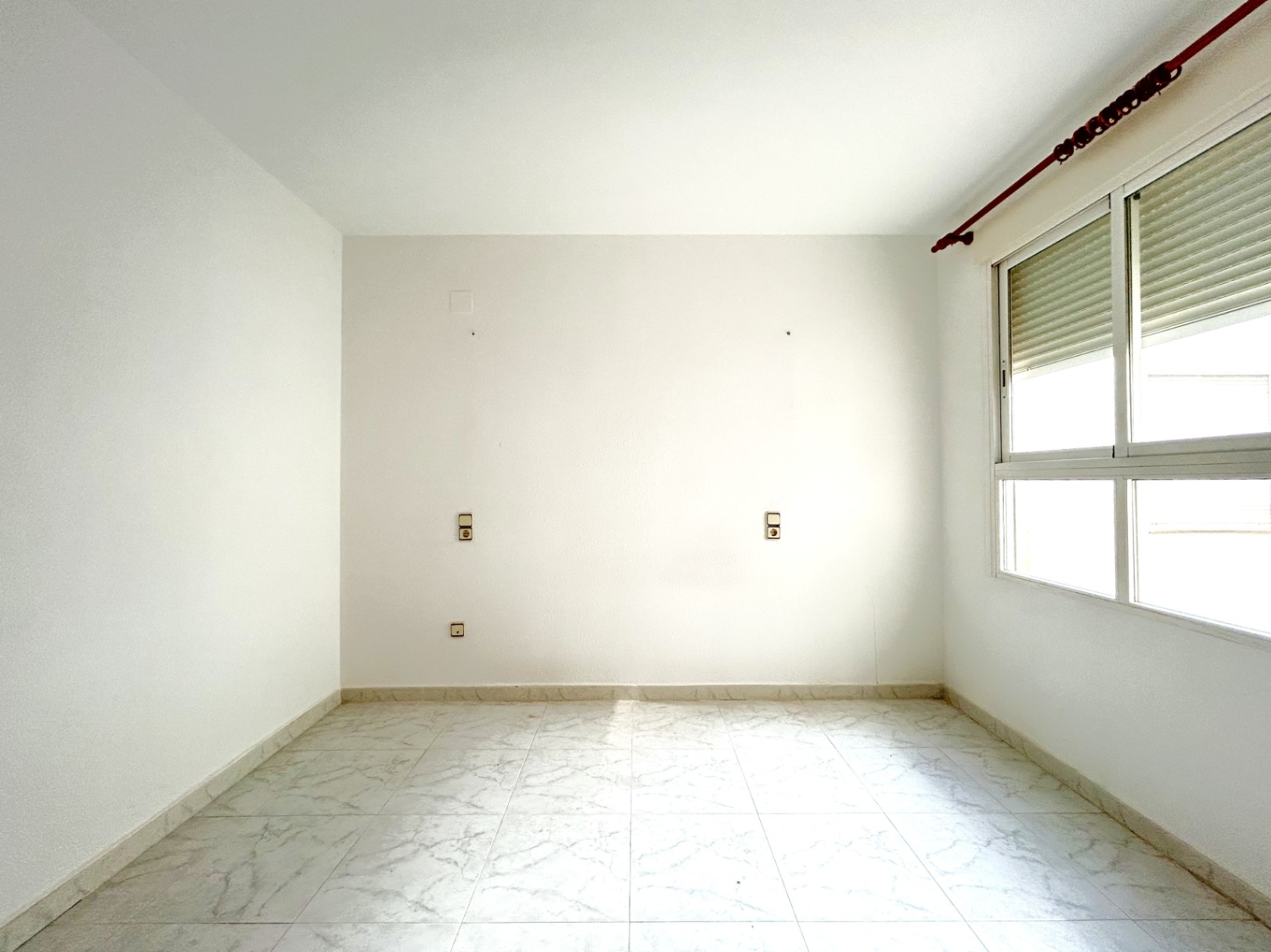Dormitorio vacío con suelos de gres efecto mármol, paredes blancas y ventana de aluminio con persiana. Dispone de tomas eléctricas simétricas para la zona del cabecero.