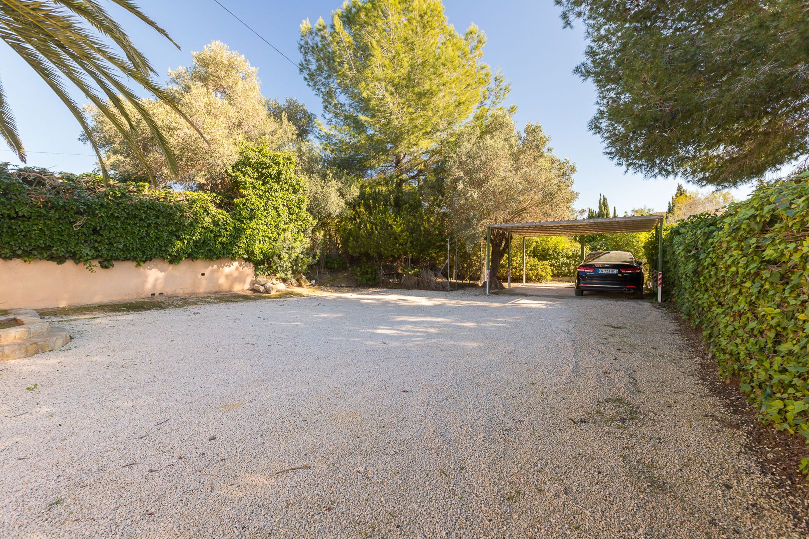 Amplia zona de aparcamiento exterior con suelo de grava, rodeada de vegetación madura y un carport techado para la protección de vehículos.