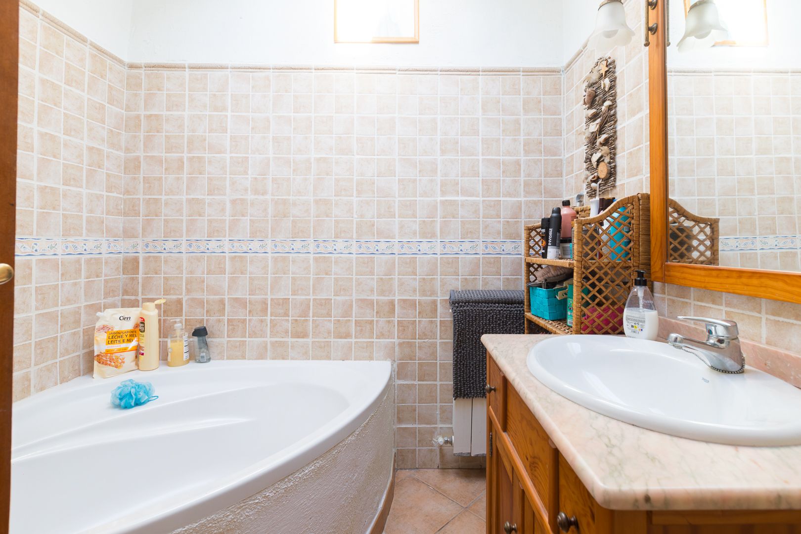 Baño con bañera de esquina, encimera de mármol sobre mueble de madera y paredes revestidas con azulejos en tonos beige y cenefa decorativa.