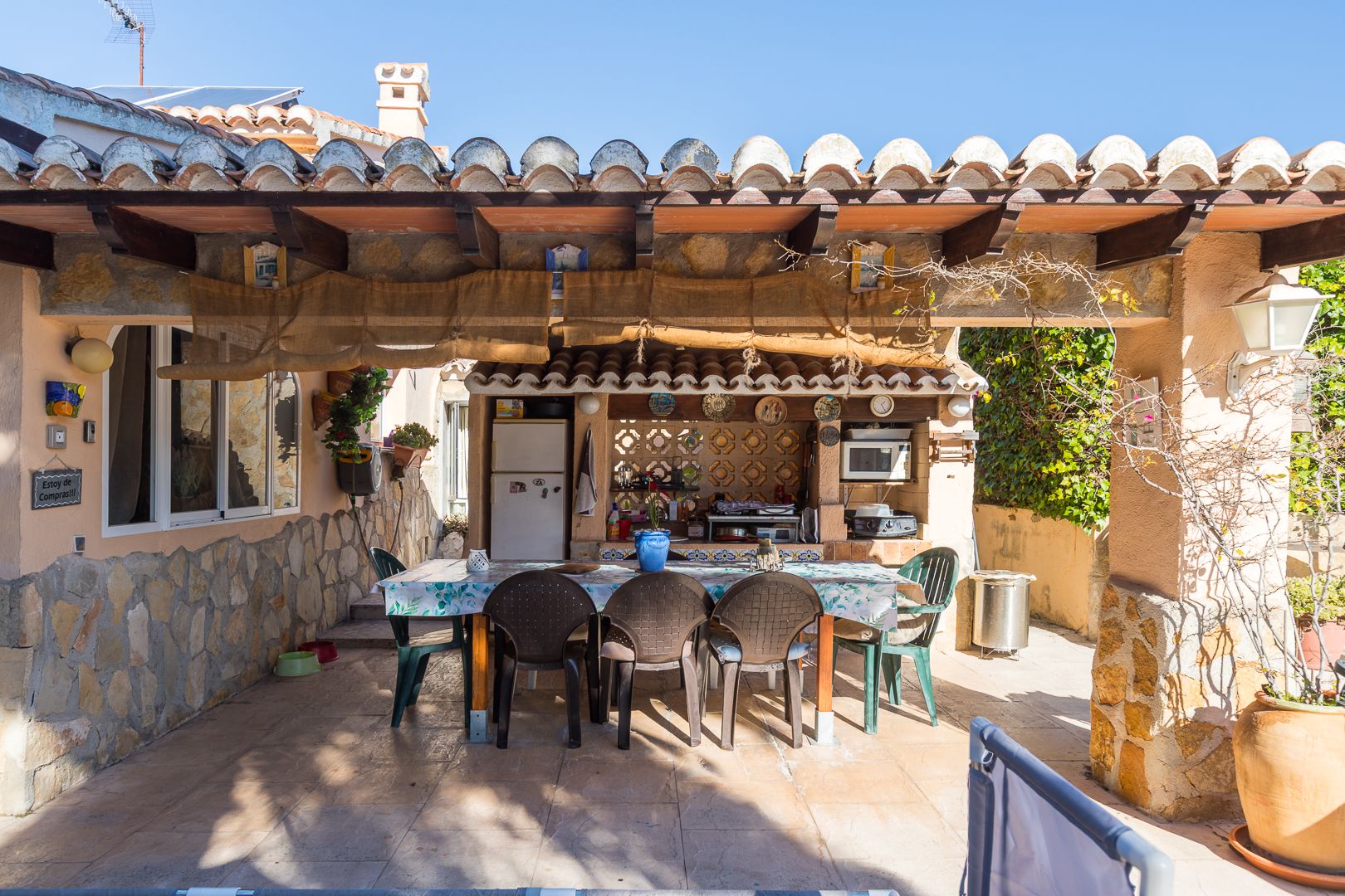 Cocina de verano y comedor exterior bajo porche de vigas de madera y teja, con paredes de piedra natural y zona equipada con electrodomésticos.