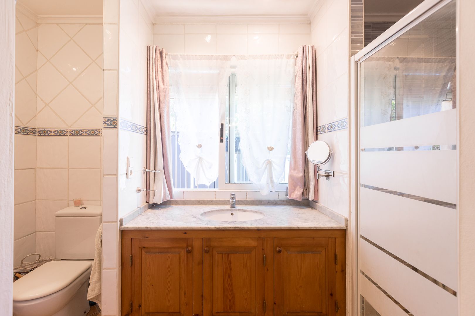 Cuarto de baño con mueble de madera, encimera de mármol y ventana frontal. Incluye sanitarios blancos, mampara de ducha serigrafiada y azulejos con cenefa decorativa.