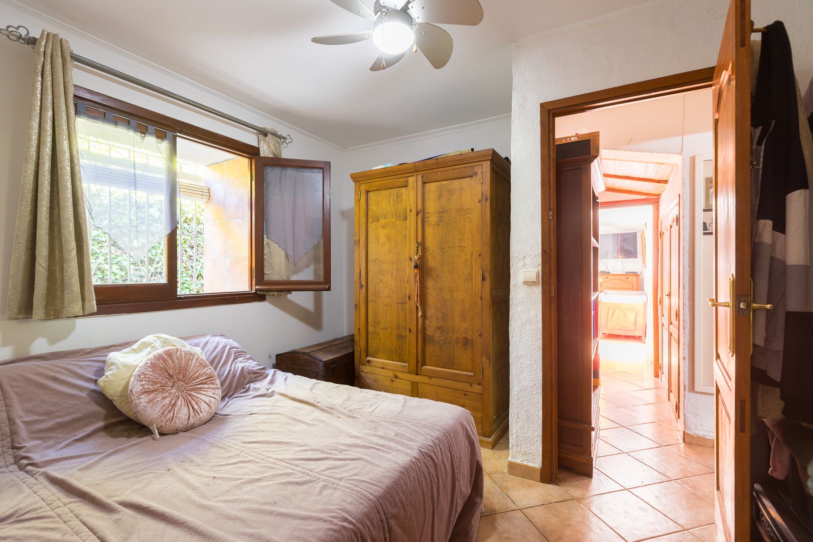 Dormitorio acogedor con armario de madera maciza, suelos de gres y ventana con marco de madera. Incluye ventilador de techo y luz natural en un ambiente funcional.