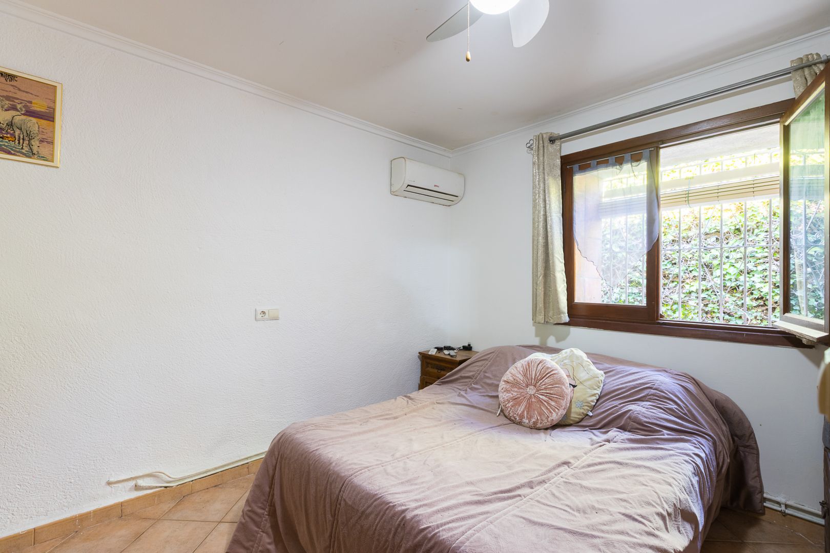 Dormitorio luminoso con aire acondicionado split, ventilador de techo y ventana con marco de madera que ofrece vistas al jardín.