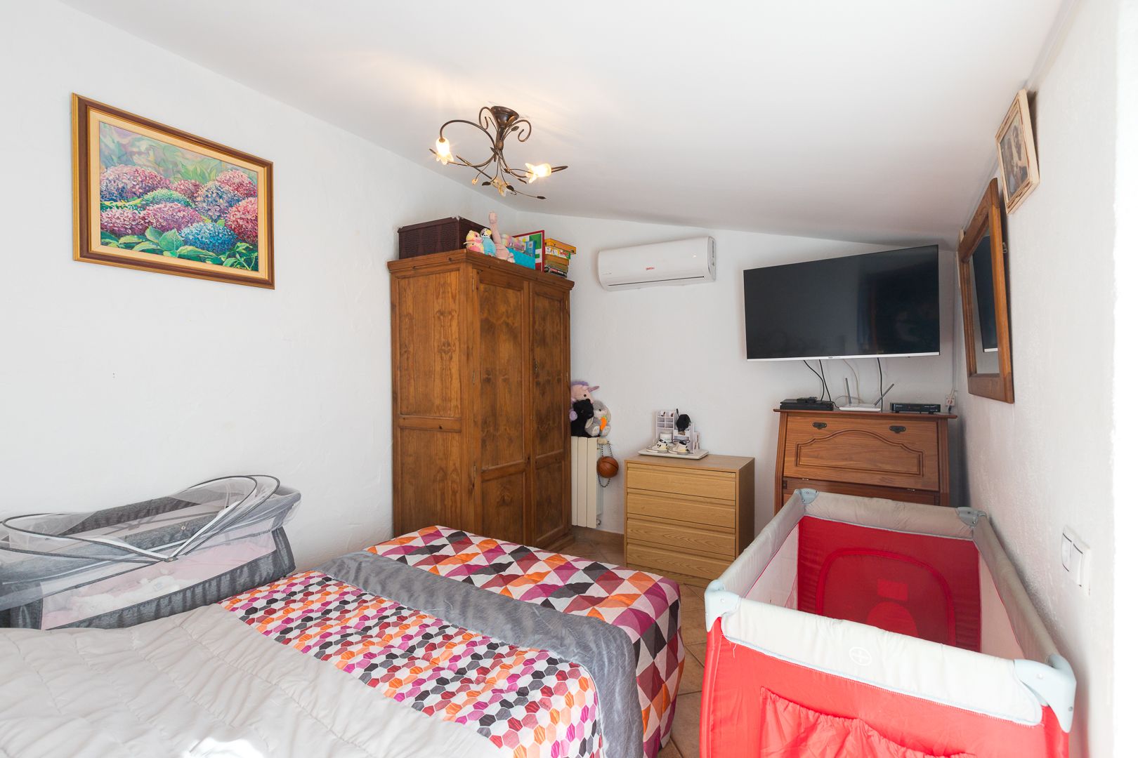 Dormitorio luminoso con techos inclinados, equipado con aire acondicionado, mobiliario de madera clásica y espacio funcional para cunas.