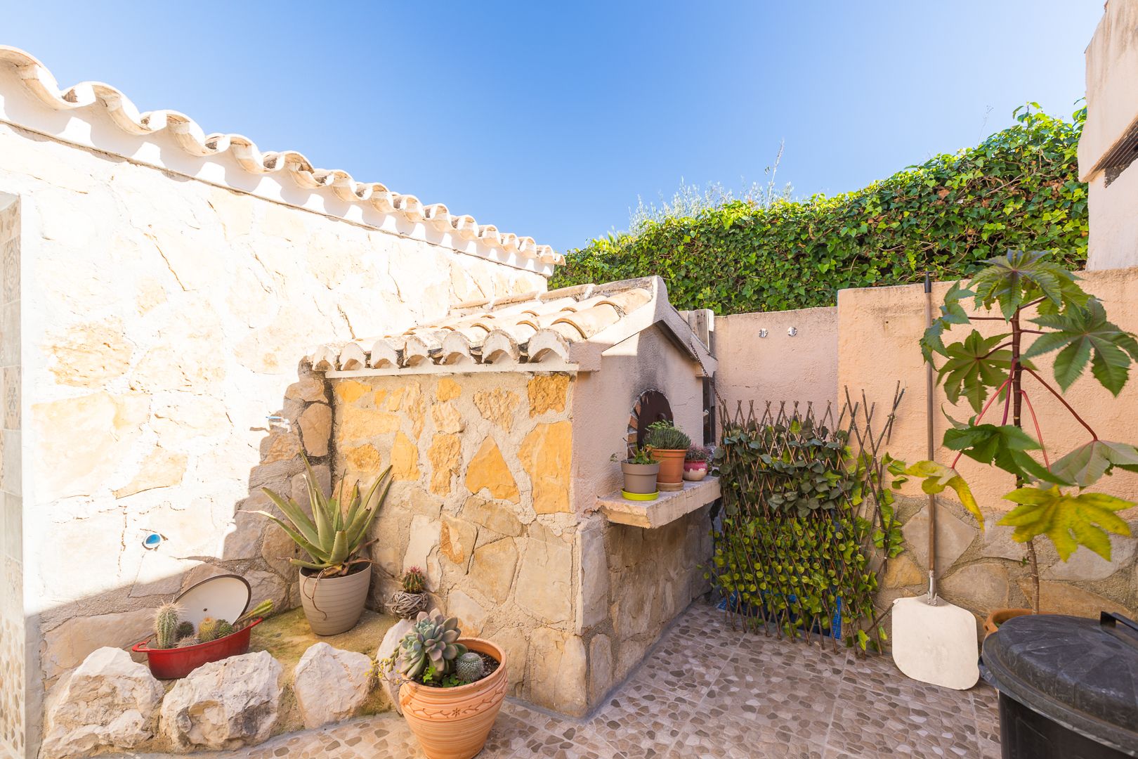 Horno de leña tradicional revestido en piedra natural con tejado de teja cerámica, situado en un patio exterior con plantas suculentas y suelos de gres.